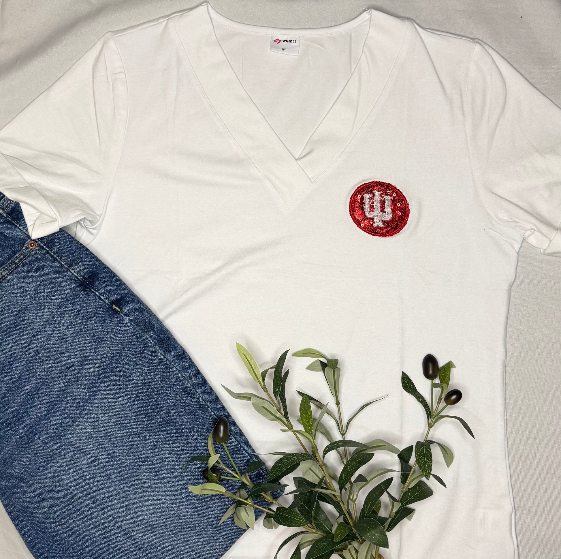 White V-neck shirt with a red and white IU trident logo, next to blue jeans and greenery.